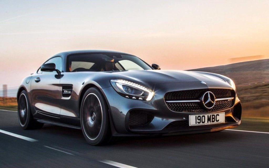 Dark grey Mercedes-AMG GT S sports coupe driving fast on a scenic road at sunset.