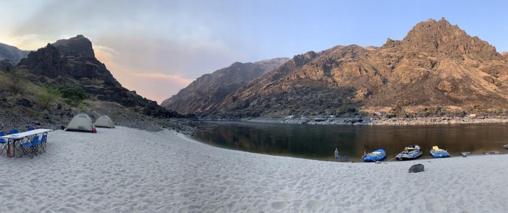 Panoramic view of a river campsite with tents and rafts on a large sandy beach surrounded by rugged canyons.