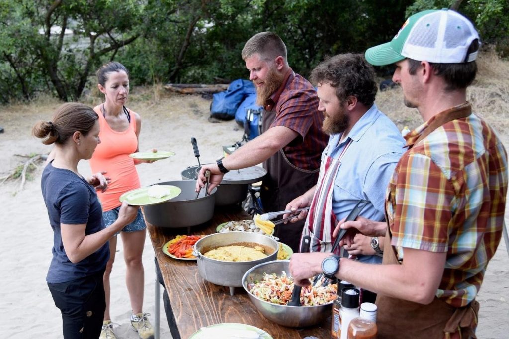Outdoor caterers serving cornbread and coleslaw to two women at a campsite meal table.