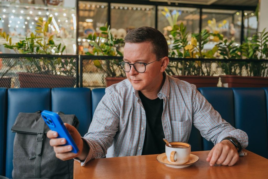 Man in glasses checking a blue smartphone while seated at a cafe booth with a cup of coffee.