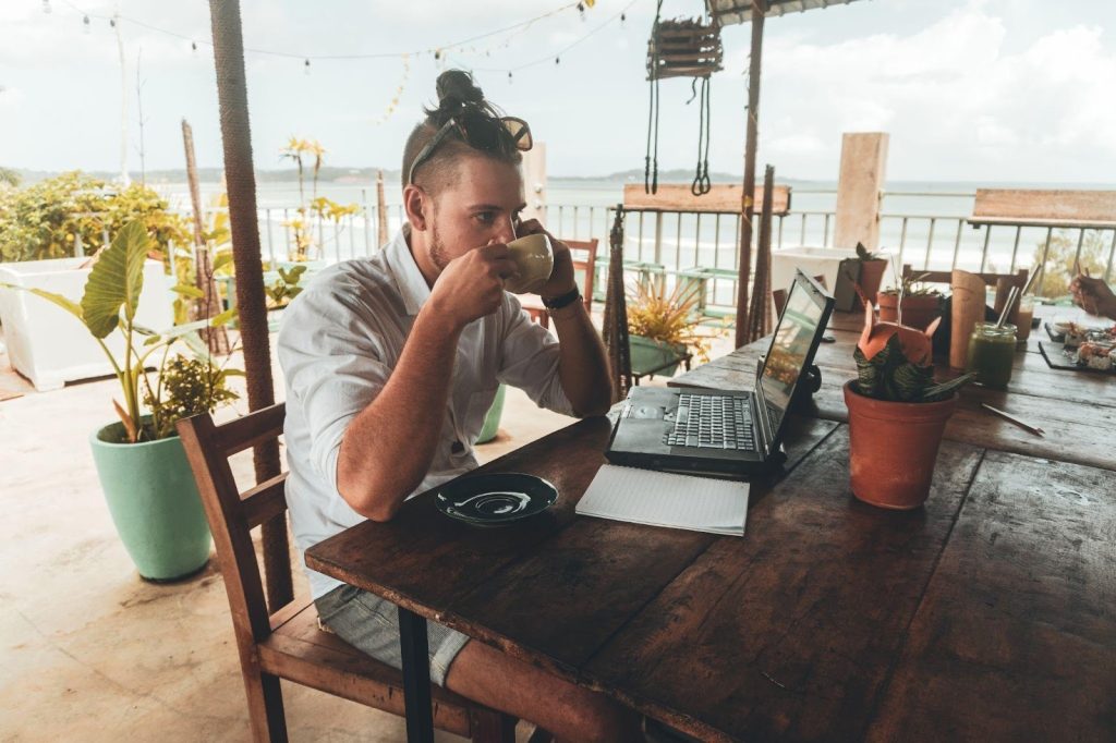 Digital nomad drinking coffee and working on a laptop at a rustic outdoor table overlooking the ocean.