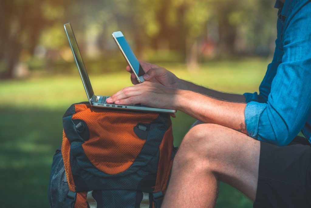 Man working outdoors using a small laptop and white smartphone, seated on an orange backpack.
