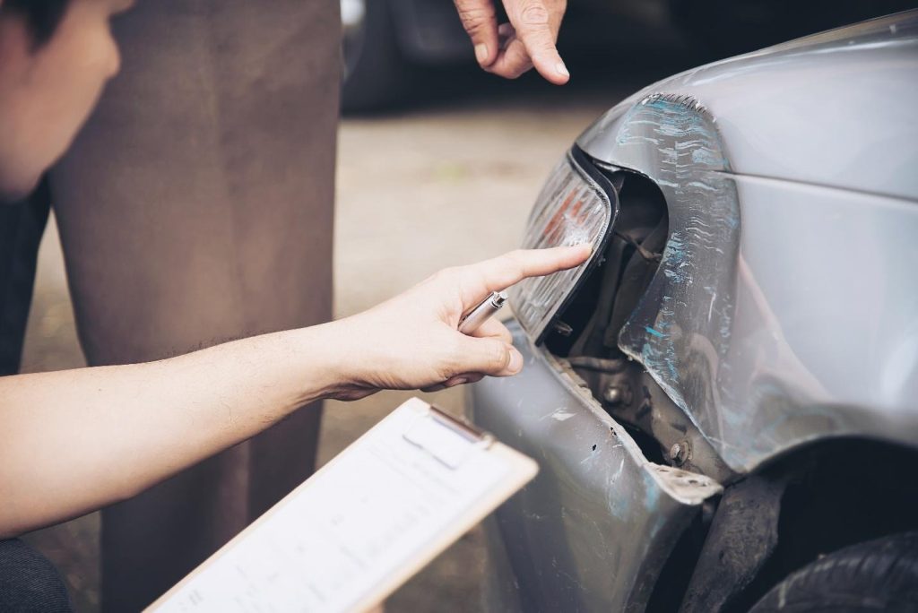 Insurance adjuster documenting damage to a silver car's headlight assembly with a clipboard and pen.