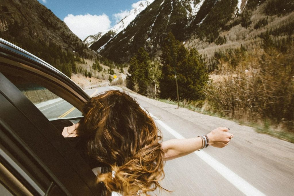 Woman with windswept hair leaning out of a car window on a scenic mountain road with snow-capped peaks.