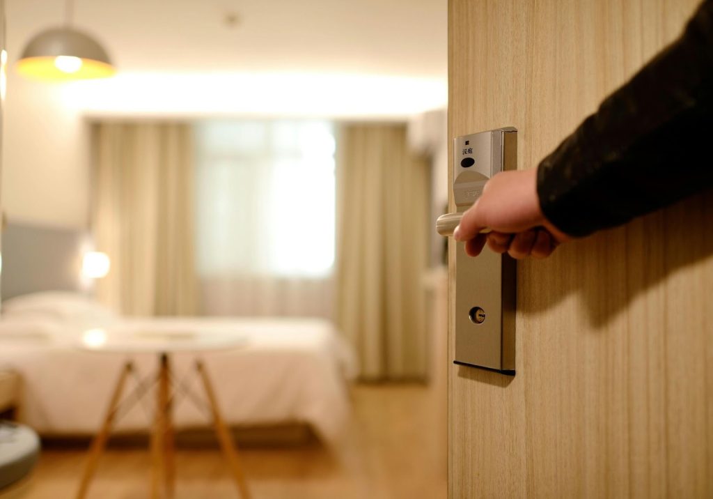 Hand using key card handle to open light wood door to a blurred, bright hotel room.