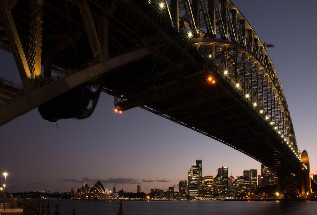 Low-angle night view of Sydney Harbour Bridge lit up with the Opera House and city skyline in the background.