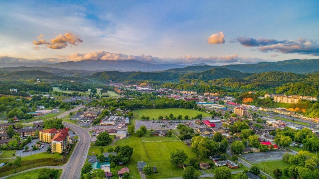 Aerial sunset view of a valley town with red-roofed buildings nestled against misty, rolling blue mountains.