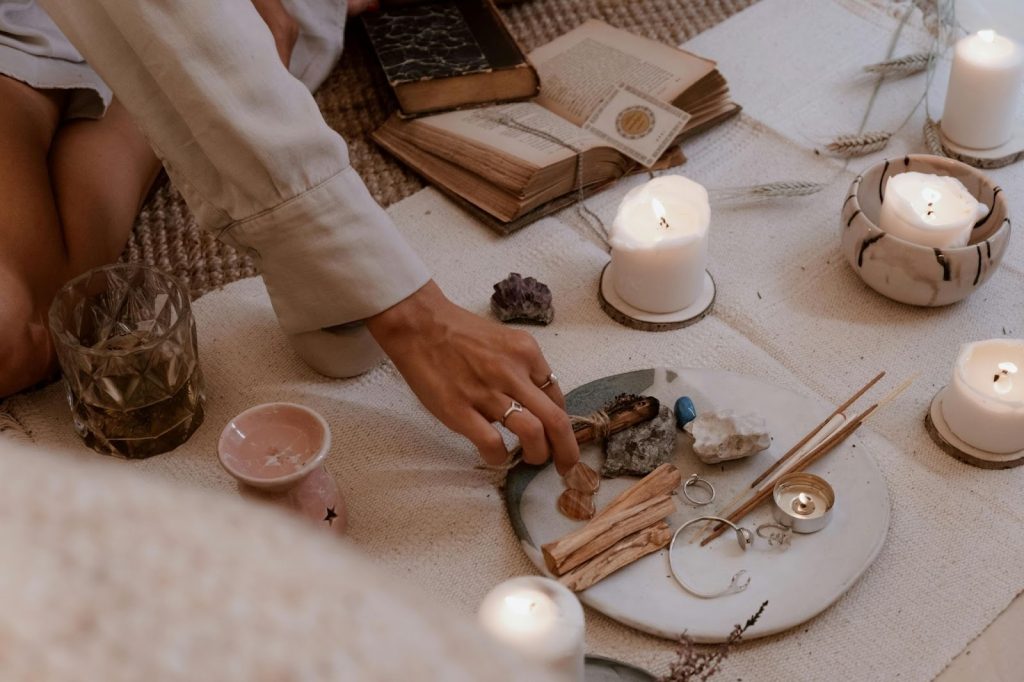 Person reaching for crystals and palo santo on a ritual plate surrounded by glowing candles and old books.