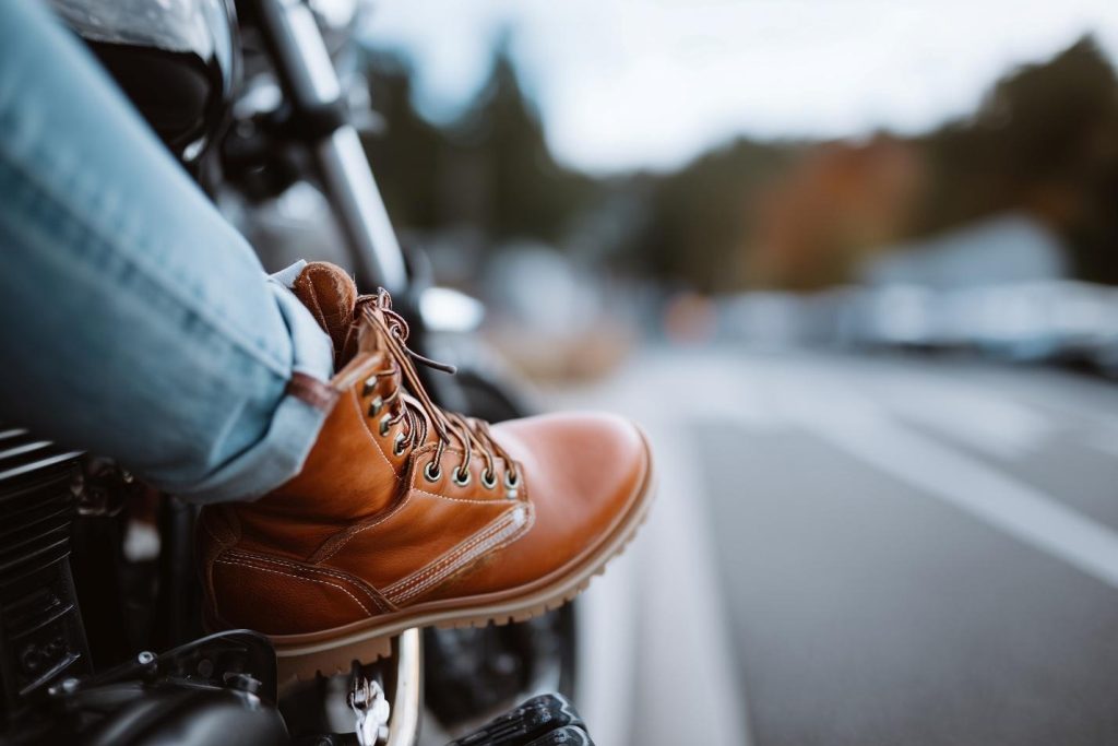 Close-up of a tan leather work boot on a motorcycle footpeg with a blurred highway road in the background.