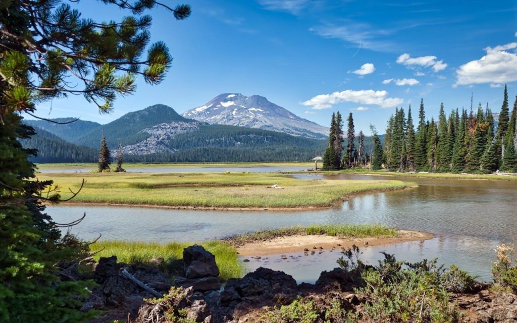 A winding river through green marshland with a snow-capped volcanic mountain under a clear blue sky in Oregon.