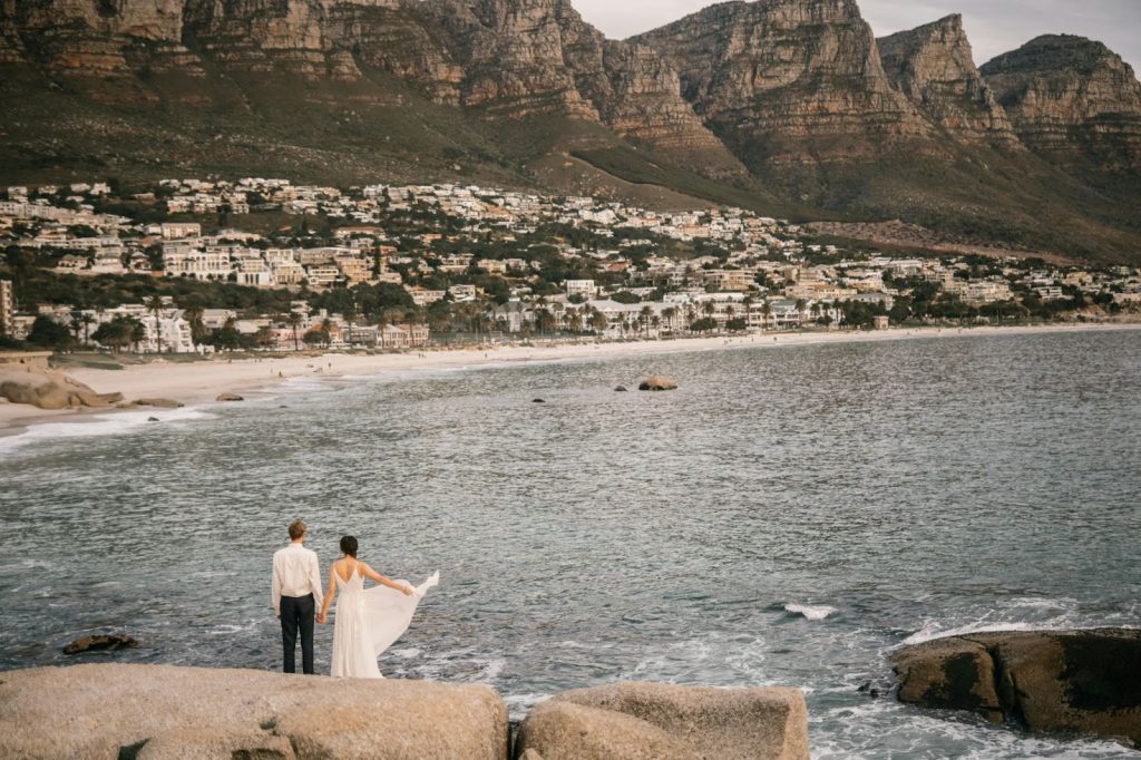 Couple standing on coastal rocks overlooking a beach town and rugged mountains at dusk.