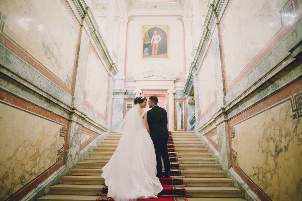 Bride and groom standing on a grand marble staircase with a red carpet in an elegant, historic palace.