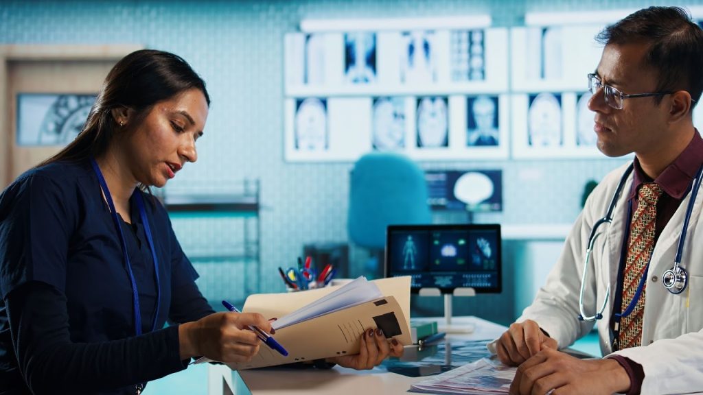 A female nurse in blue scrubs and a male doctor in a white coat review a patient file in a high-tech medical lab.