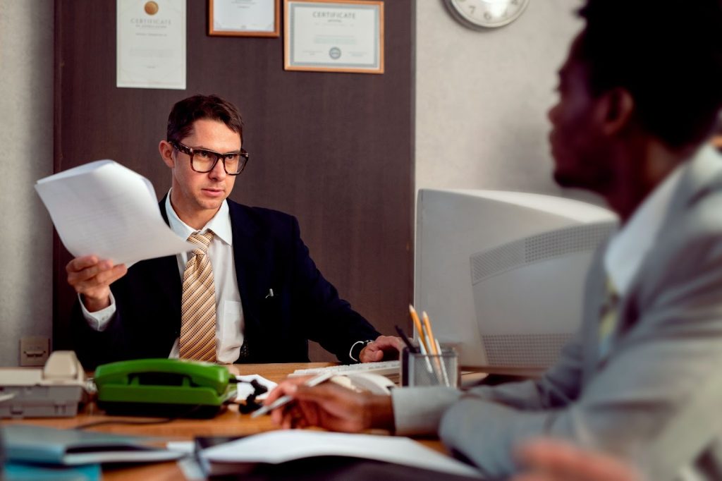 A man in a suit holds up papers while speaking to a colleague in a retro office with a green phone and beige monitor.