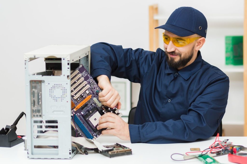IT Technician in blue uniform and safety glasses installing a purple motherboard into a desktop computer casing.