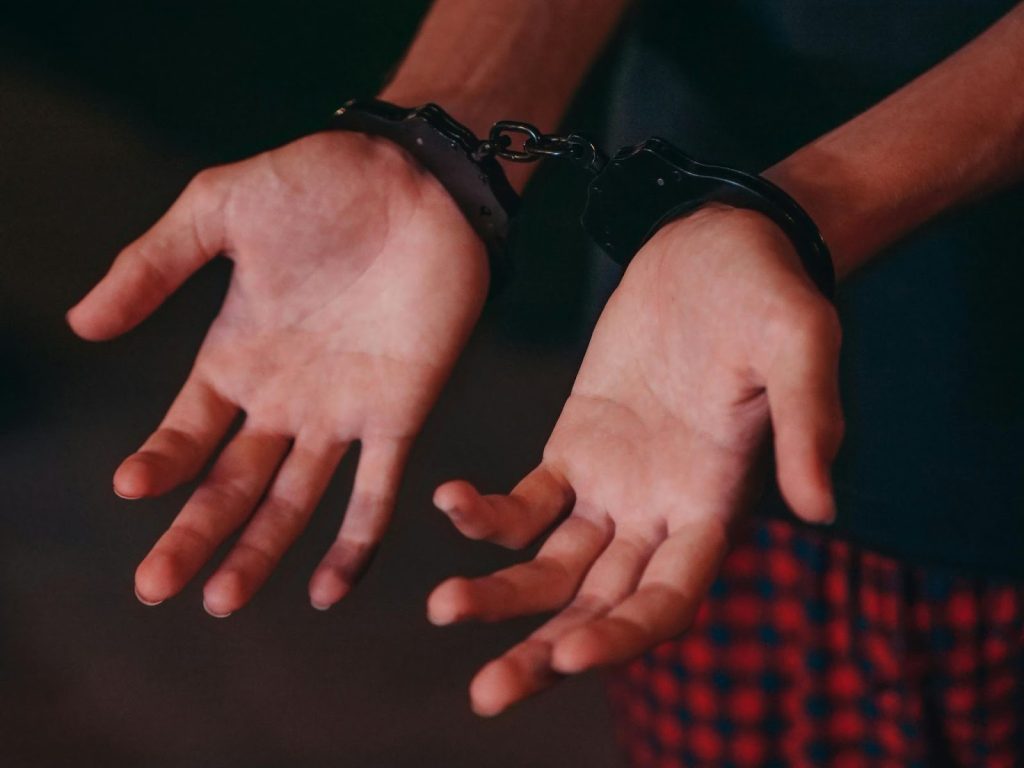 Close-up of a person's hands held out in black metal handcuffs against a dark, moody background.