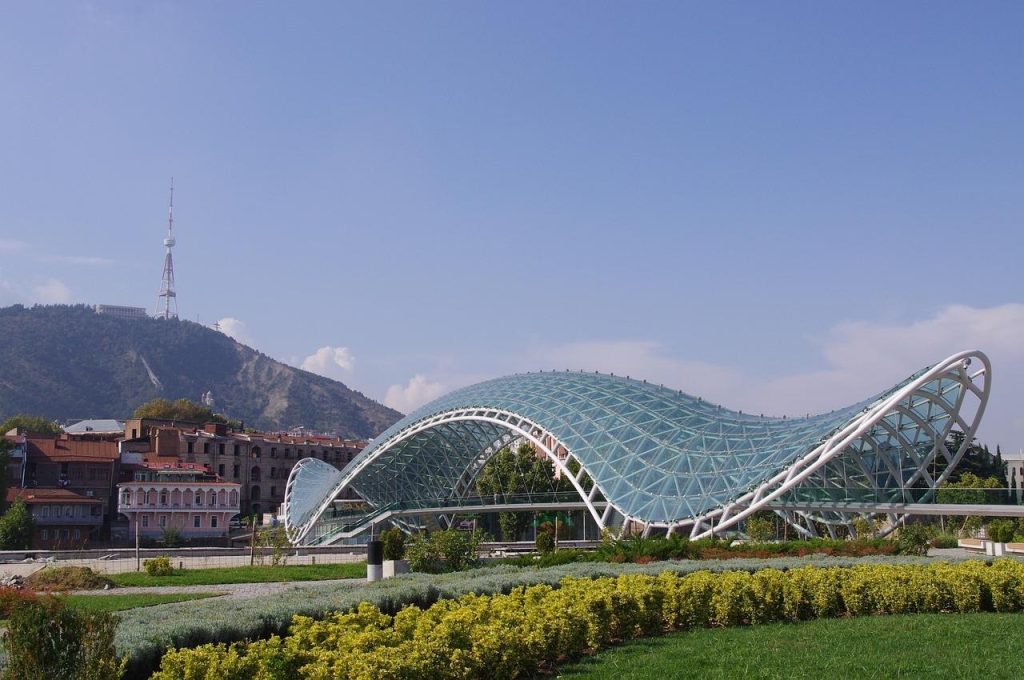 Modern glass Bridge of Peace in Tbilisi, Georgia, with Mtatsminda Mountain and TV tower in the background.