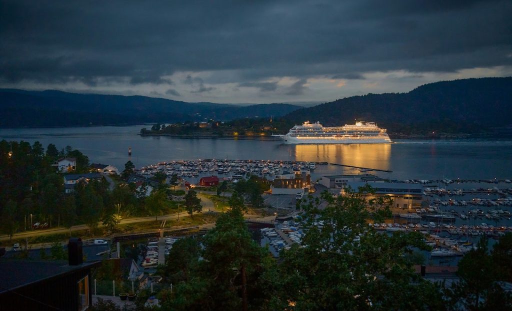 Illuminated cruise ship reflecting in a harbor at dusk with a marina and forested hills in the background.