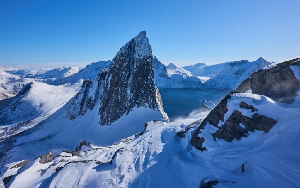 Snow-covered Segla mountain peak towering over a deep blue fjord in Senja, Norway, under a clear sky.