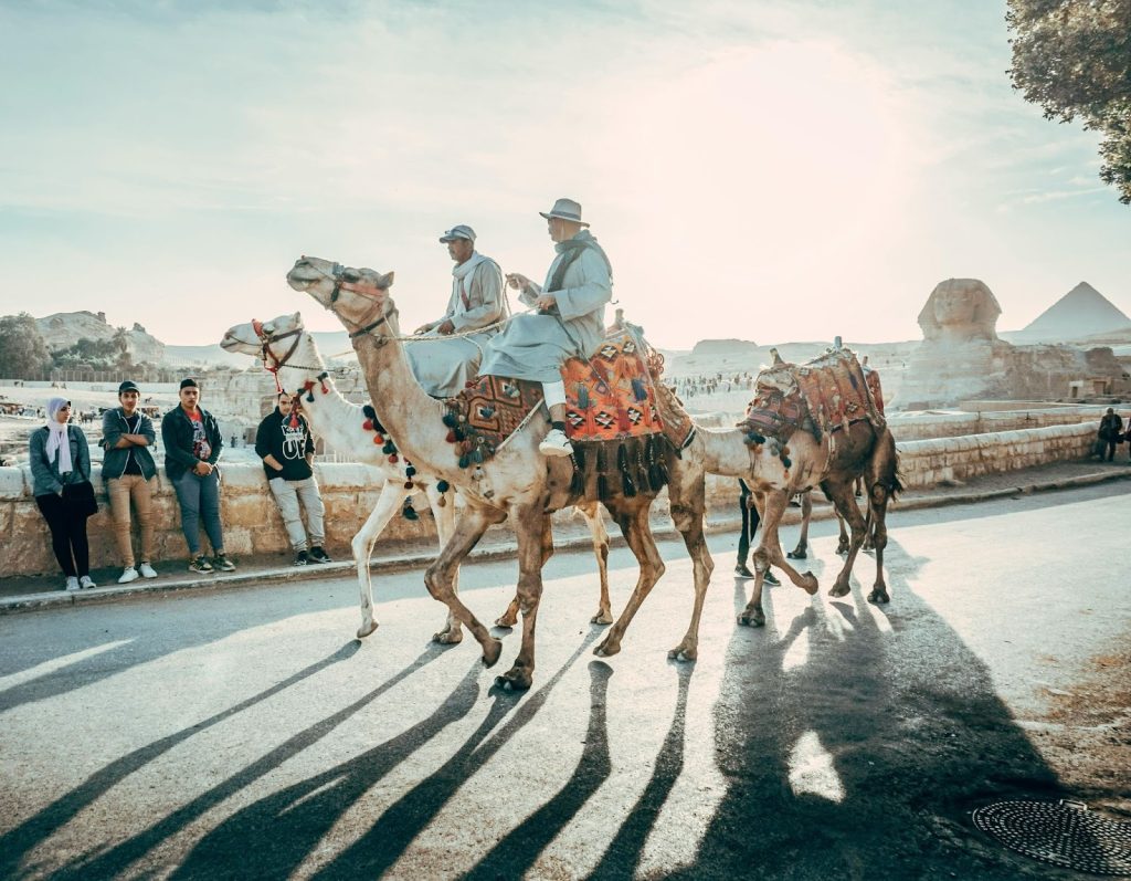 Men riding camels on a road at the Giza Plateau with the Great Sphinx and Pyramids in the hazy background.