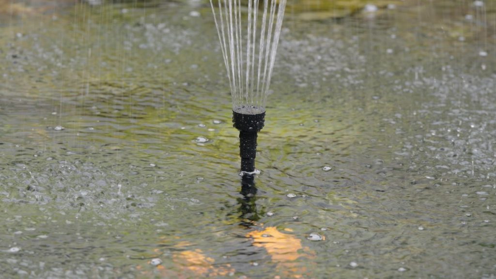 Close-up of a black pond fountain nozzle spraying water streams onto a rippling garden pond surface.