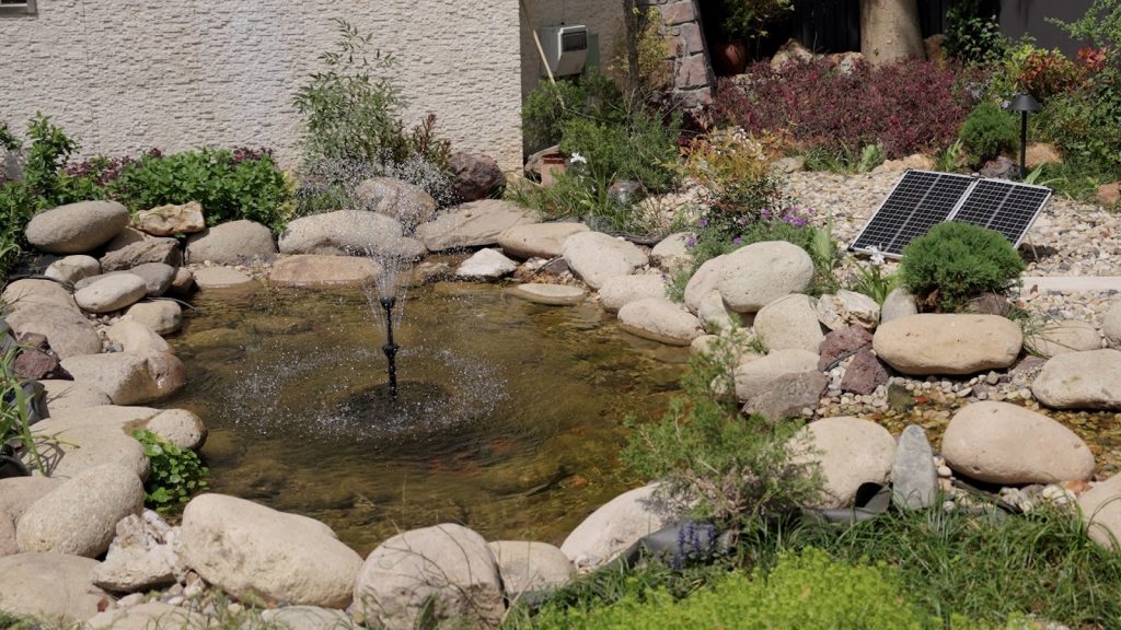 Solar powered pond fountain with a multi-tier spray head and solar panel in a stone-lined backyard garden pond.