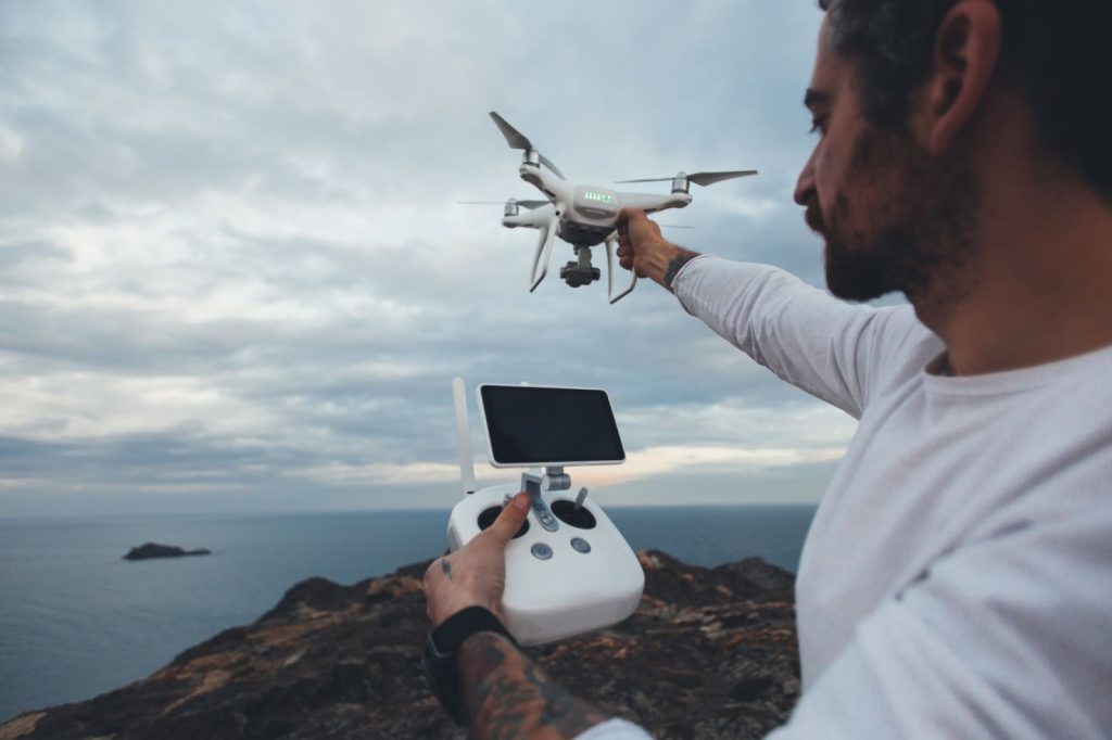 Man with tattoos holding a white drone and remote controller on a rocky cliff overlooking the ocean.