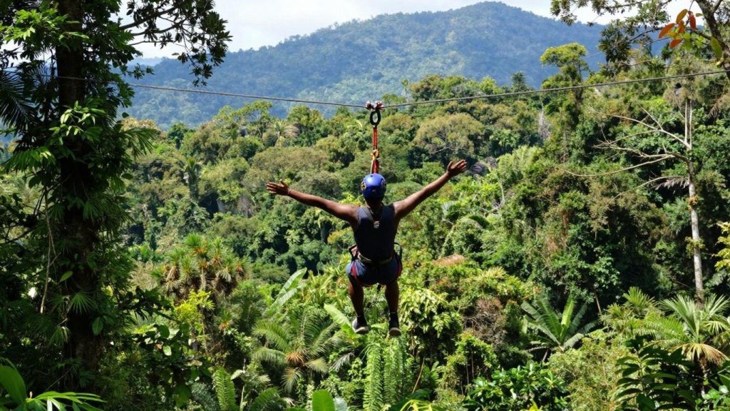 Person ziplining over a lush tropical rainforest canopy with arms outstretched against a mountain backdrop.