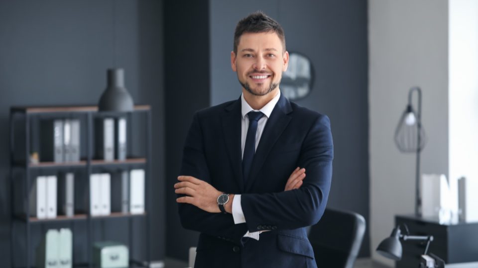 Professional man in a navy suit smiling with arms crossed in a modern, minimalist office.