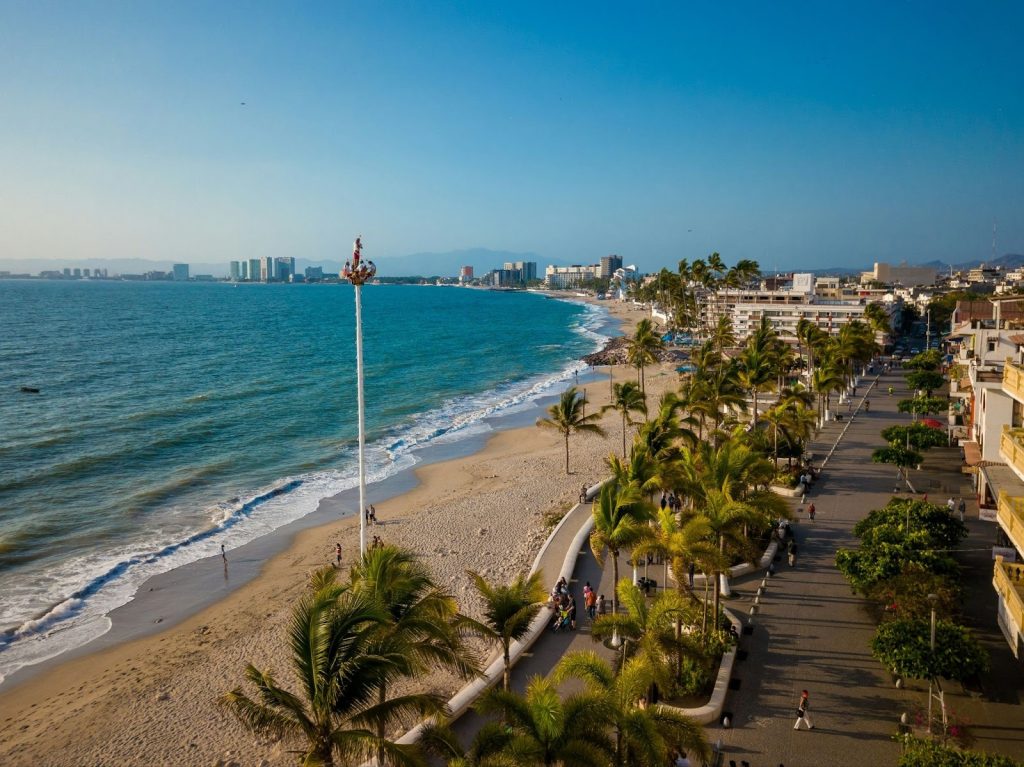 Aerial view of a sunny tropical beach and palm-lined boardwalk with distant city high-rises under a clear blue sky in Puerto Vallarta