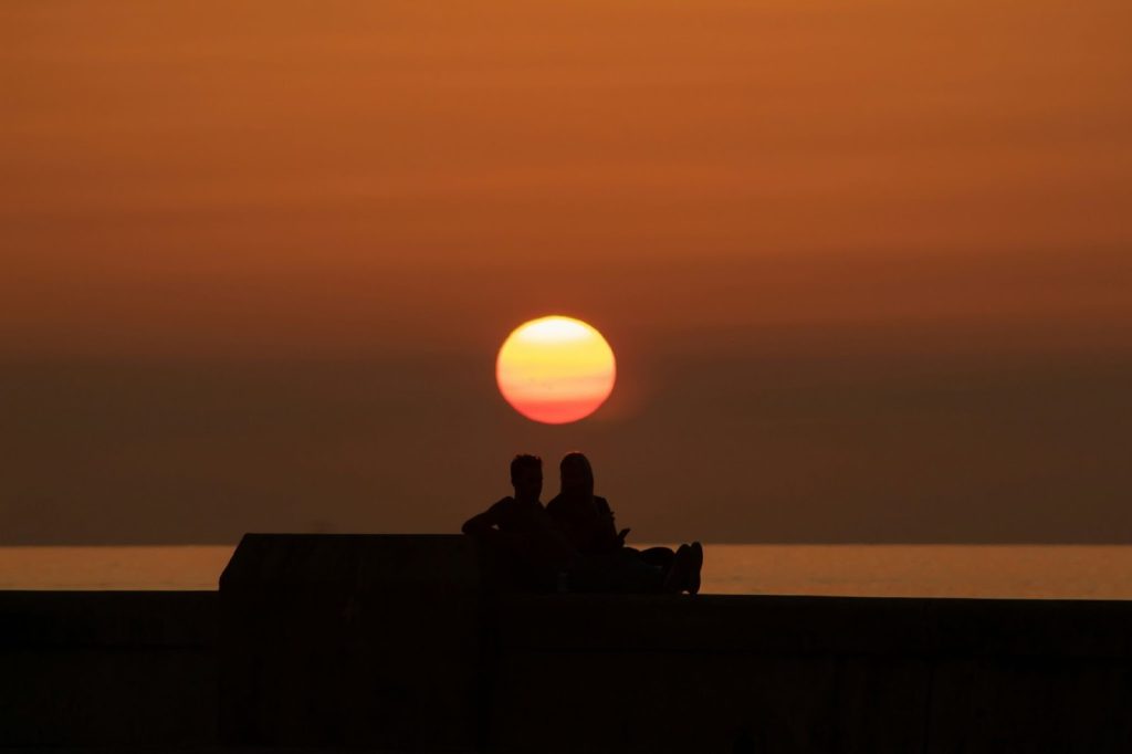 Silhouette of a couple sitting by the ocean against a massive, glowing orange sun at sunset.