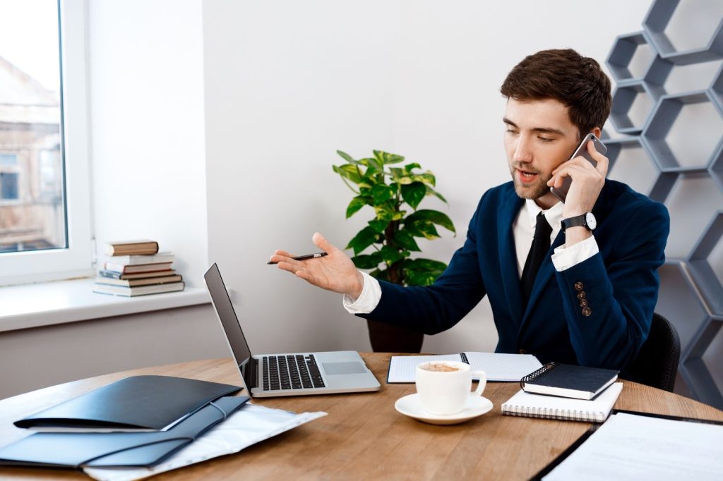 Businessman in a blue blazer talking on a phone and gesturing toward a laptop at a bright office desk.