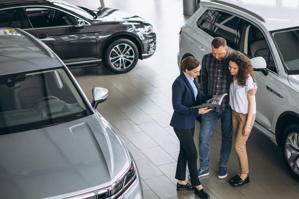 A saleswoman shows a brochure to a couple in a modern car showroom surrounded by silver and white SUVs.