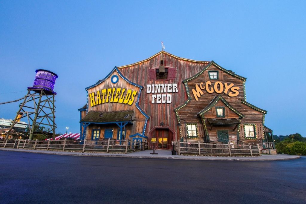 Exterior of the Hatfield & McCoy Dinner Feud theater with rustic wood siding and neon signs at twilight.