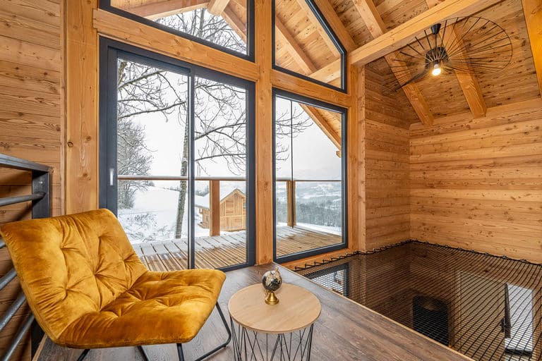 Modern log cabin mezzanine with a mustard yellow chair, floor net hammock, and large windows overlooking a snowy forest.