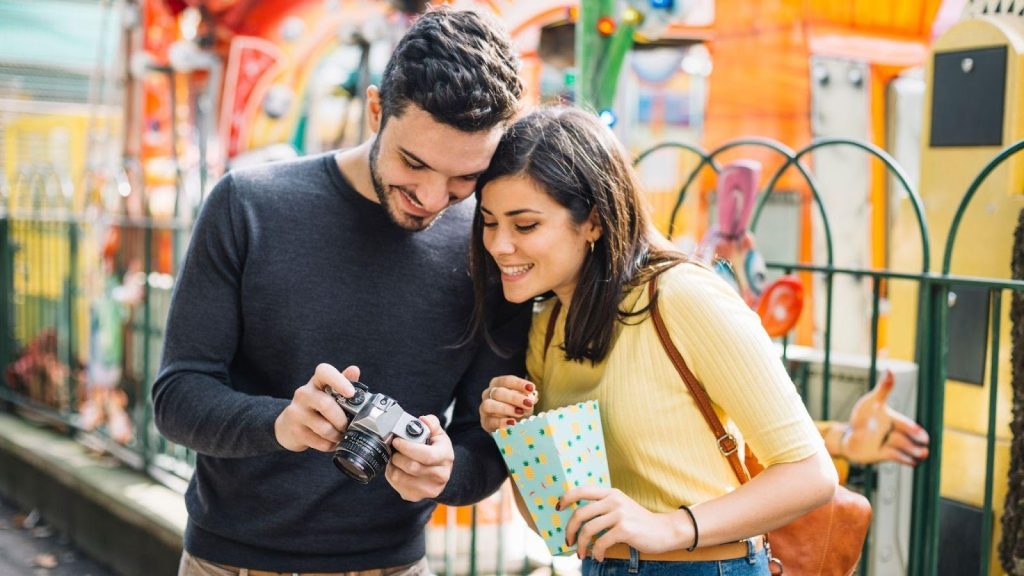 A smiling couple at a fairground looking at a vintage camera while holding a box of popcorn.