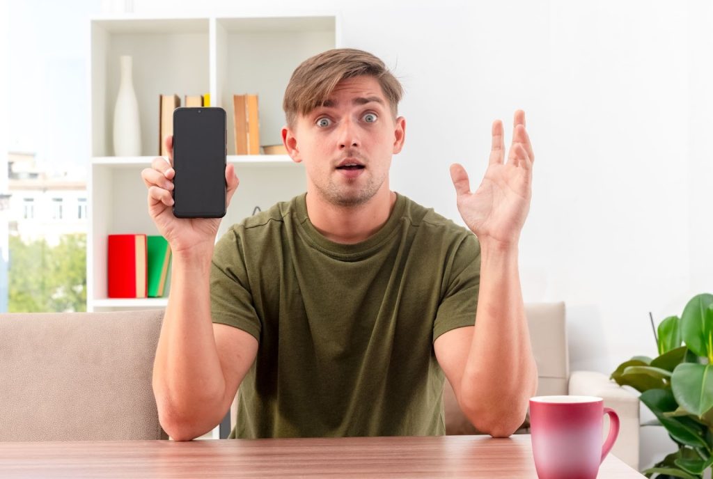 A shocked man in a green shirt holding a smartphone with a blank screen and gesturing in surprise.