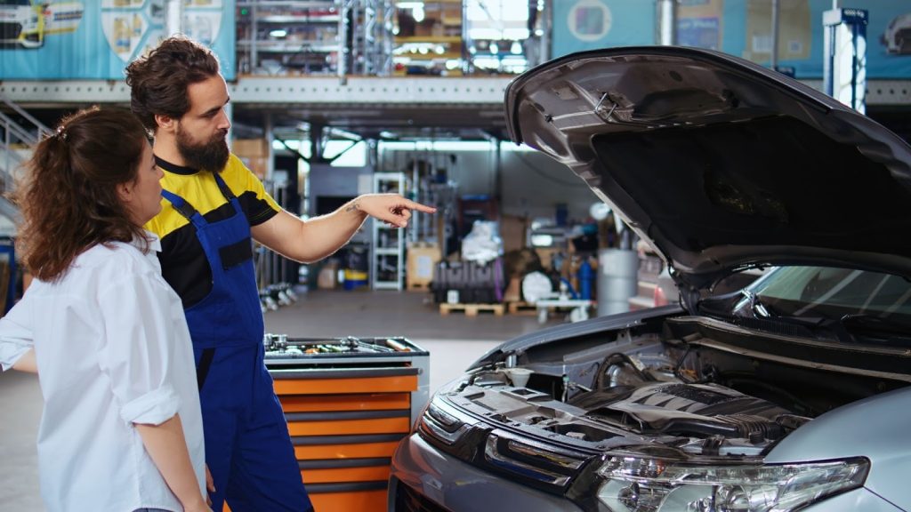 Mechanic in blue overalls pointing to a car engine while explaining repairs to a female customer in a garage.