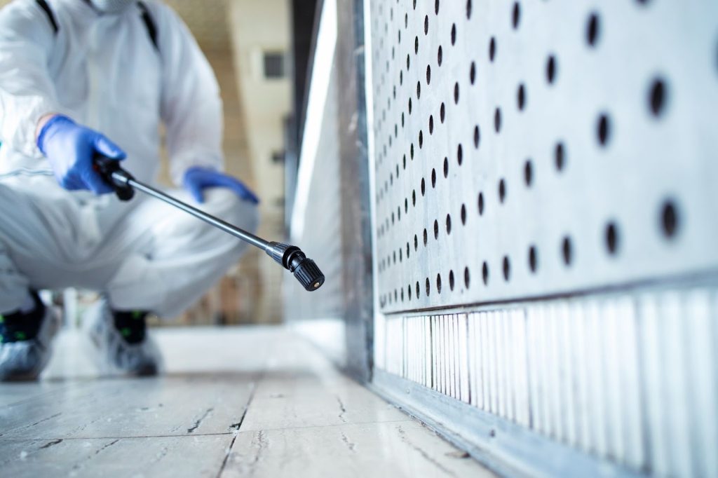 Professional in white hazmat suit and blue gloves spraying a commercial wall for pest control or sanitization.