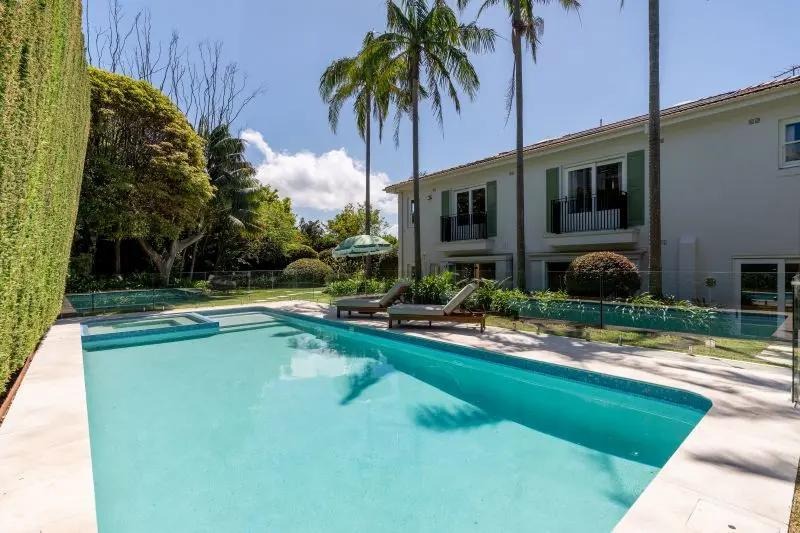 Sydney Luxury backyard featuring a turquoise swimming pool, stone patio, and palm trees in front of a modern white house in Sydney