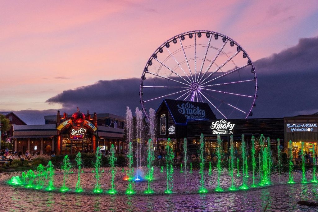 Illuminated green fountain and purple Ferris wheel at an outdoor entertainment plaza under a pink sunset sky in ole smoky
