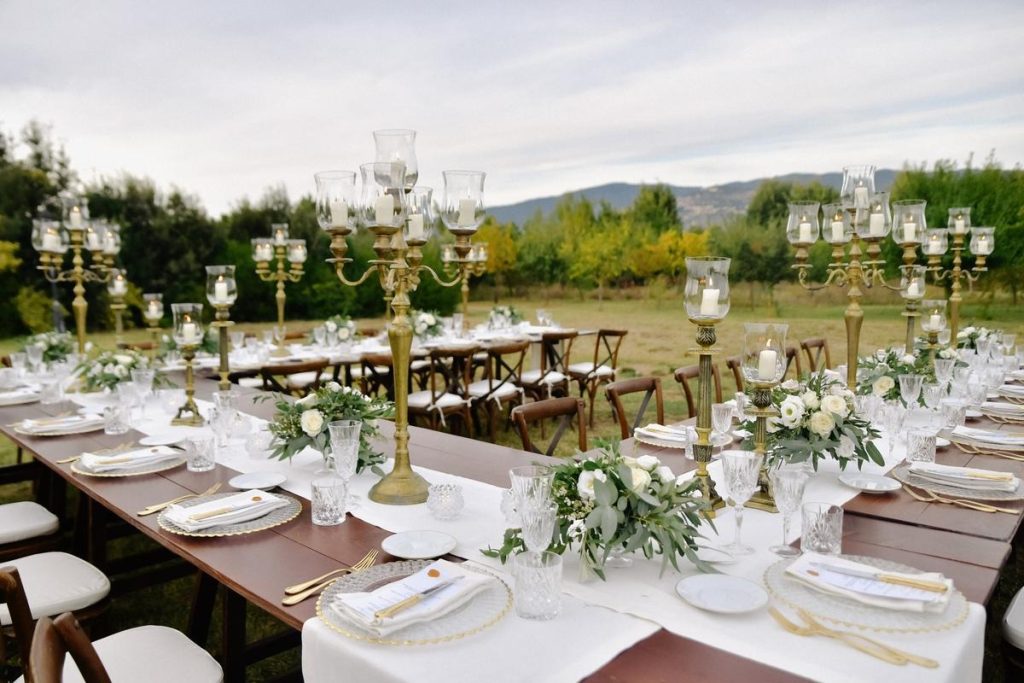 Elegant outdoor wedding reception featuring long wooden tables, tall brass candelabras, and white floral centerpieces.