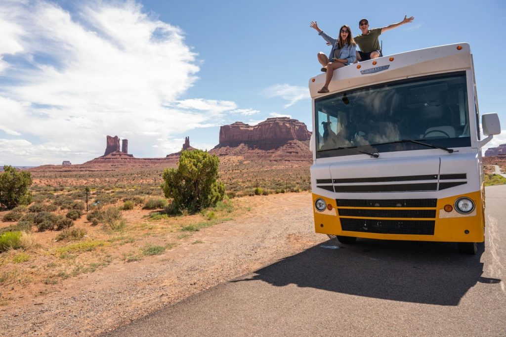 Couple cheering on top of an RV in Monument Valley, Arizona, with red rock buttes and desert landscape.