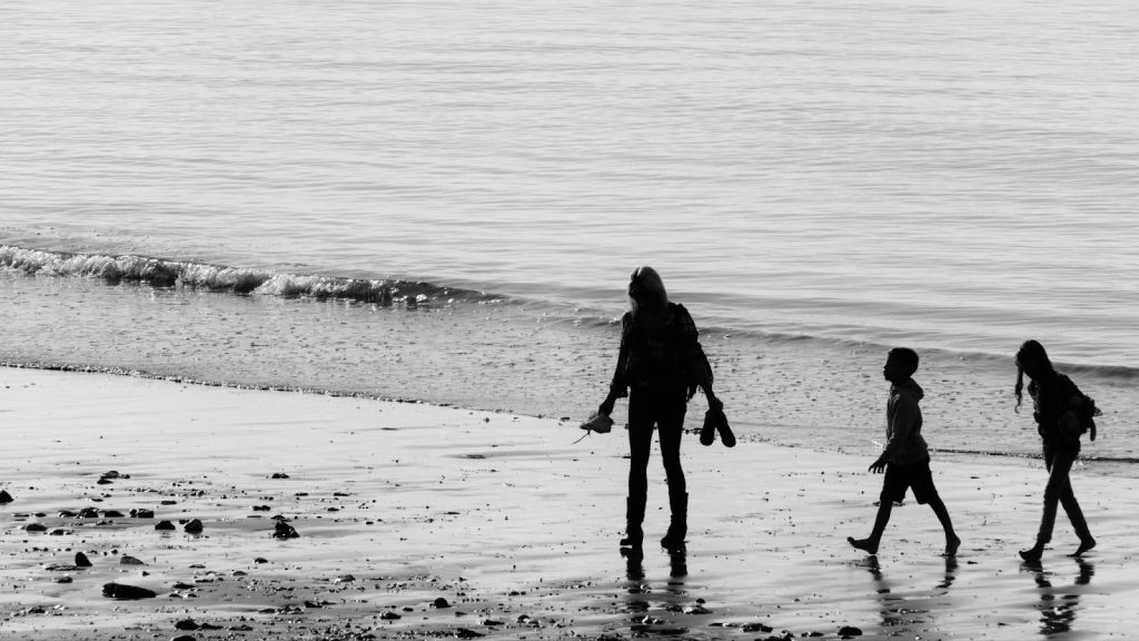 Black and white silhouette of a woman and two children walking on a reflective beach at low tide.