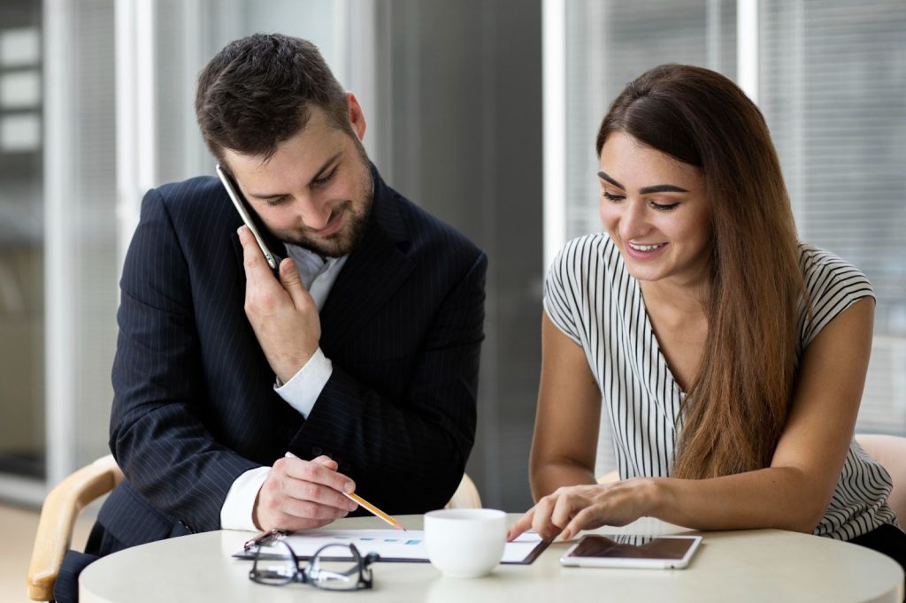 Two colleagues reviewing business charts at a table while one talks on a smartphone in a bright office.