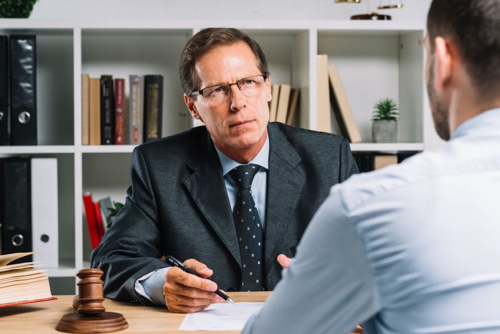 A serious lawyer in a suit and glasses discussing documents at a desk with a gavel and legal books.