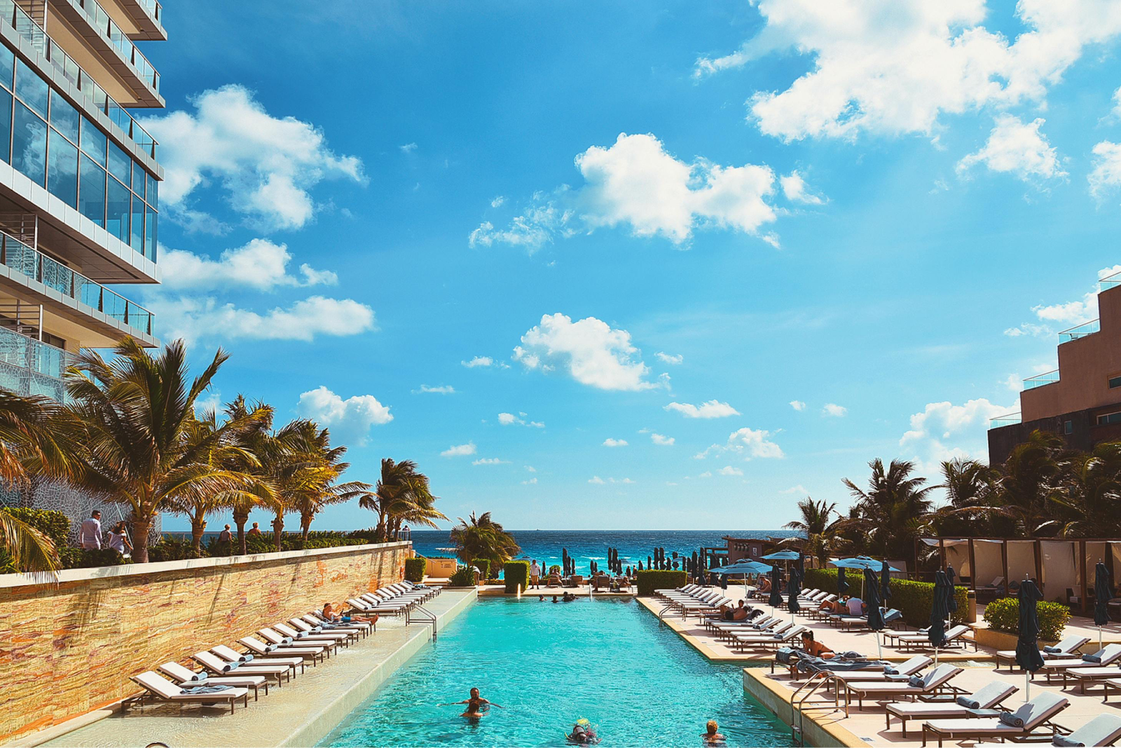 Luxury infinity pool at a modern beachfront resort with rows of lounge chairs, palm trees, and a clear blue ocean view.