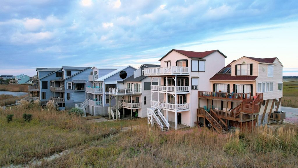 A row of colorful multi-story beach houses with wooden decks overlooking golden sea grass under a cloudy sky.