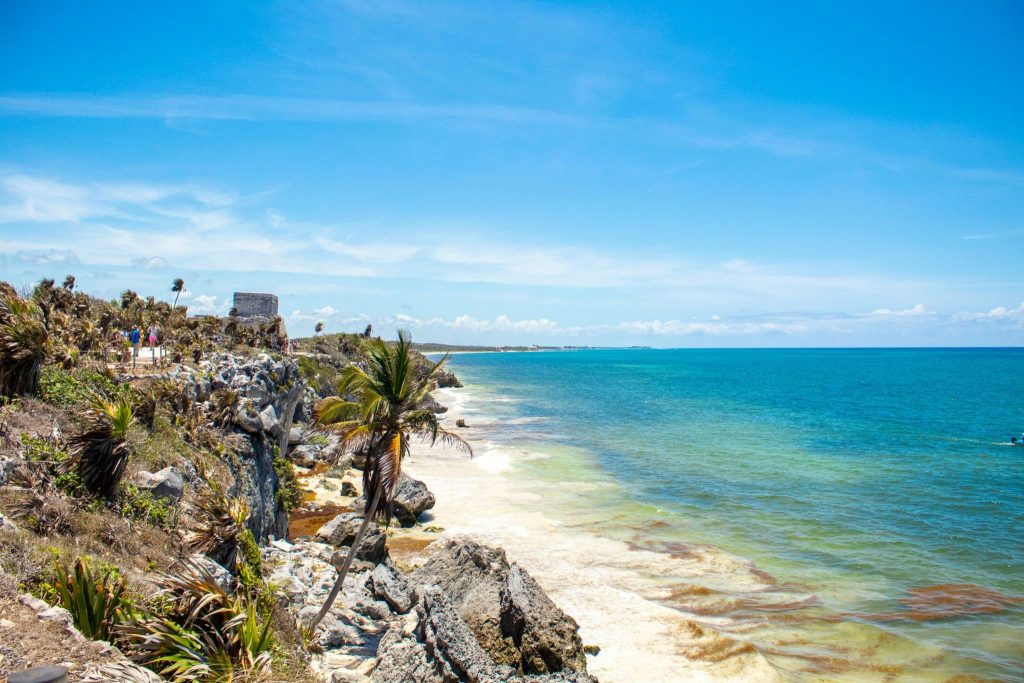 The ancient Mayan ruins of Tulum on a rocky cliff overlooking turquoise Caribbean waters under a bright blue sky.