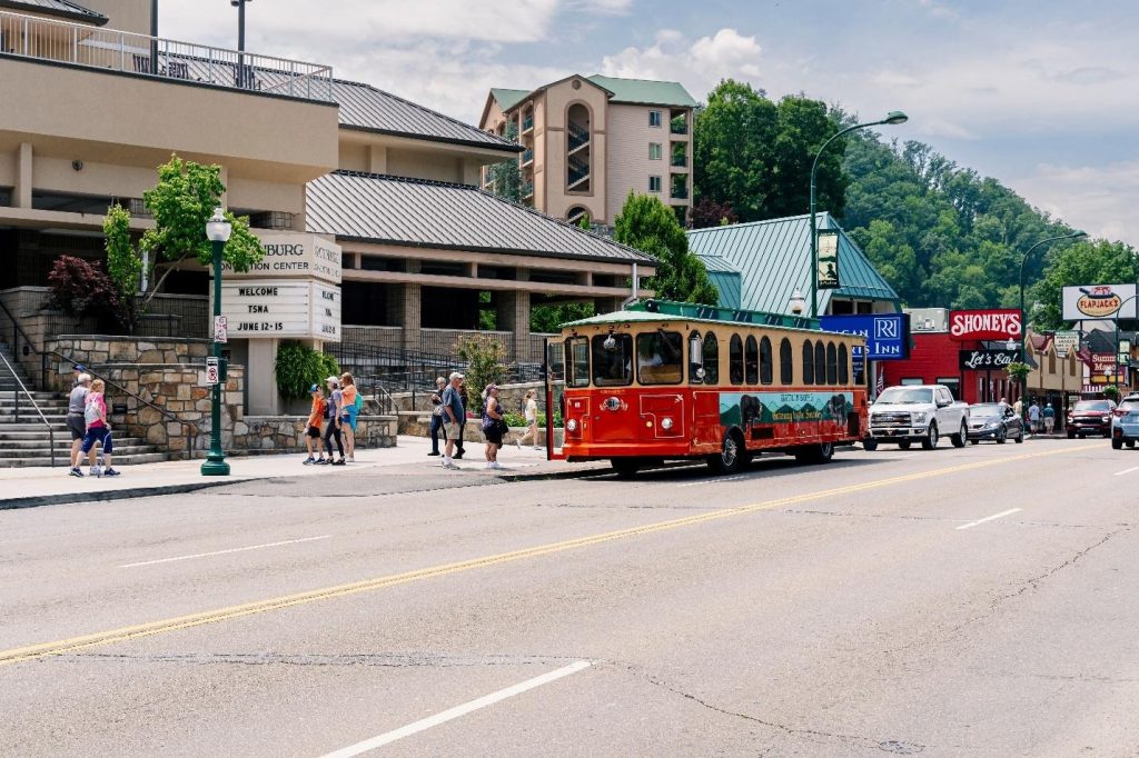 Red vintage-style tourist trolley driving past the Gatlinburg Convention Center with green mountains in the background.
